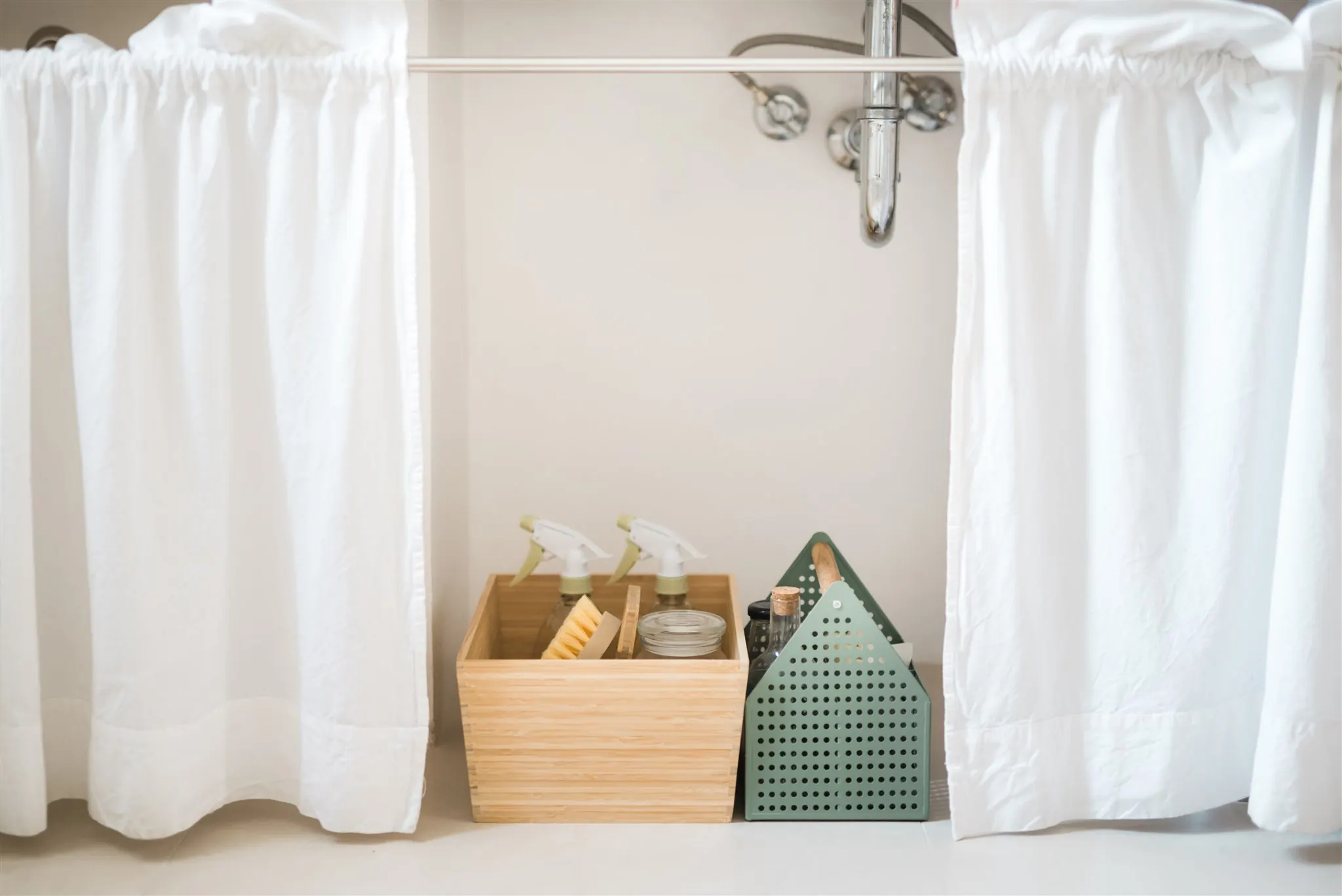 Cleaning supplies in a bamboo box and a caddy under the sink, behind a linen curtain — where household waste quietly accumulates