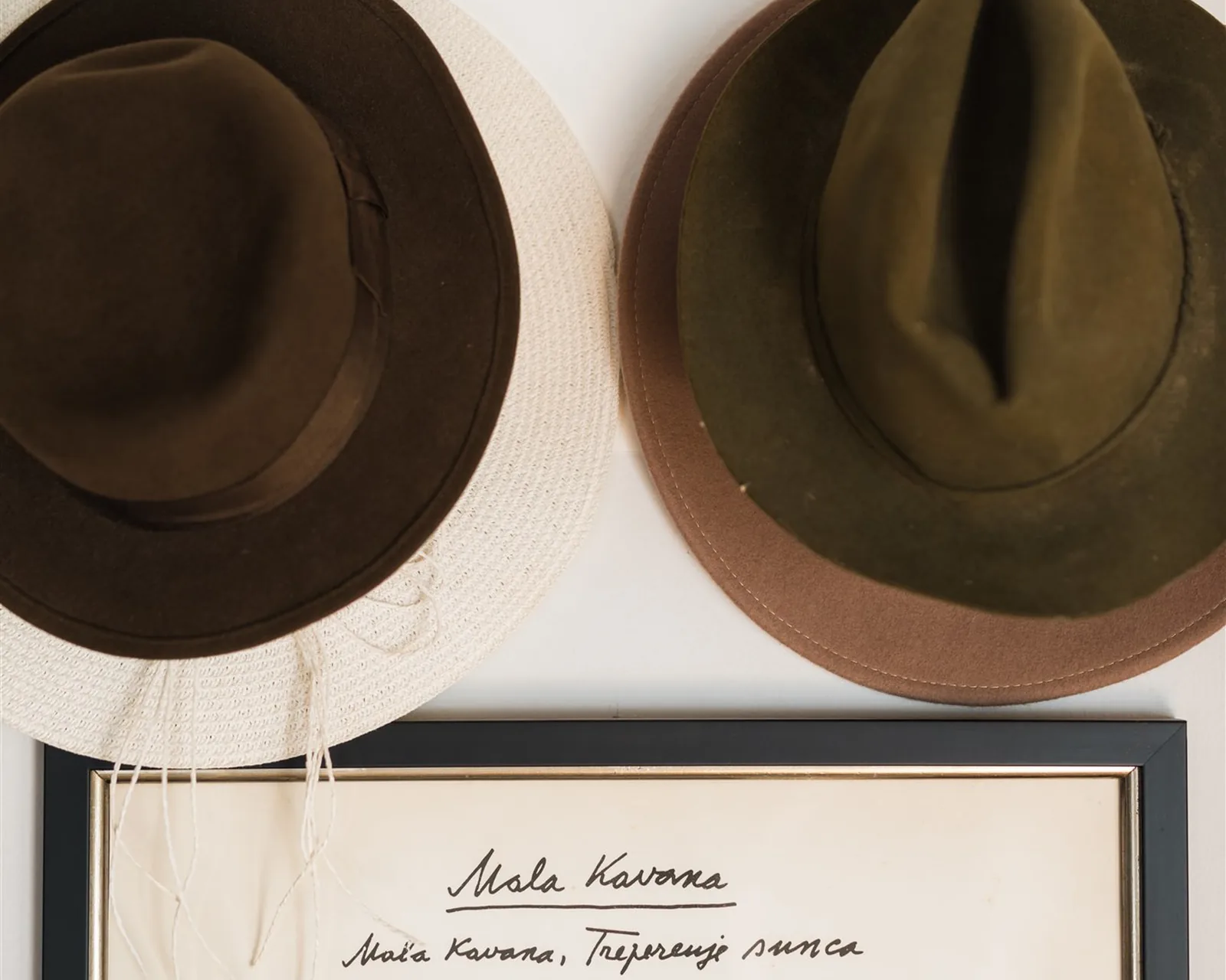 A brown and an olive hat hanging on a white wall above a framed handwritten poem — hats as wall art