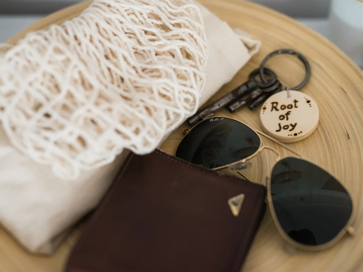 A Root of Joy key ring with keys, sunglasses and a cloth bag on a wooden tray