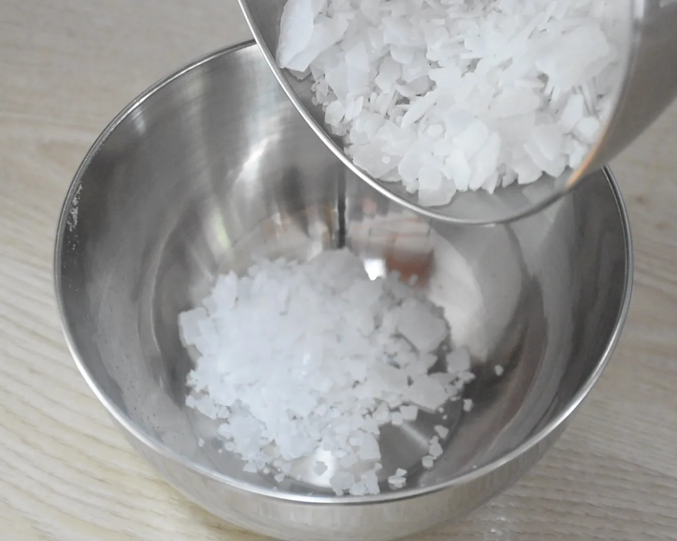Hydroxide flakes being poured from a smaller bowl into a larger stainless steel bowl