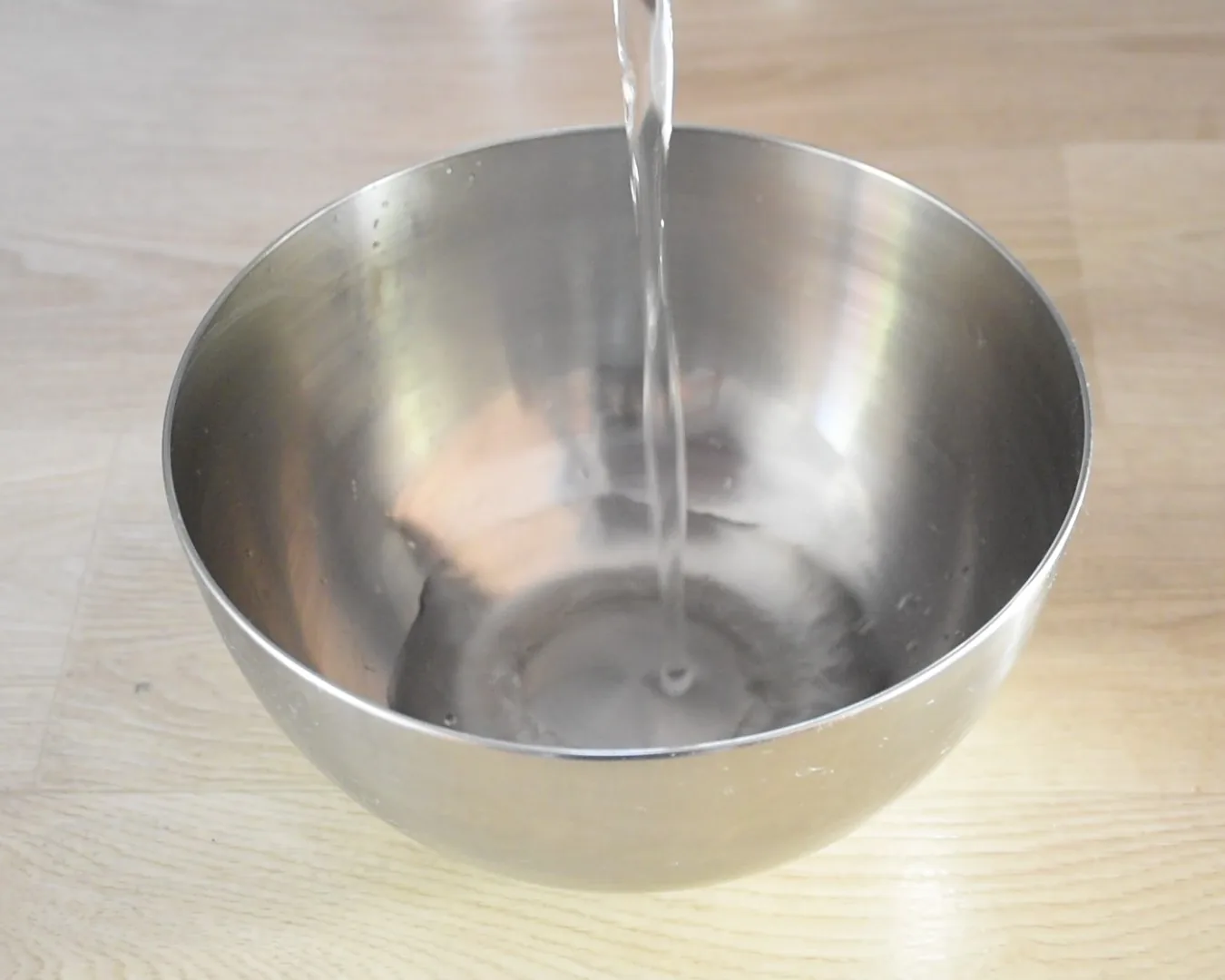 Water being poured into a stainless steel bowl on a pale wooden worktop