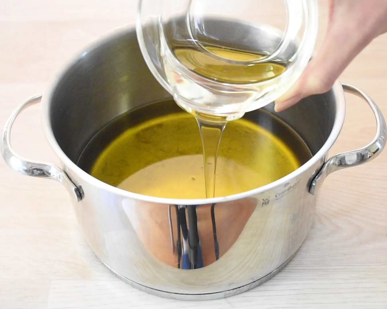 Oil being poured from a glass jug into a large stainless steel pot